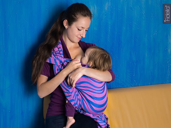 Eine Mutter stillt ihr Baby in einem Tragetuch von Fidella im Design Shebra pink